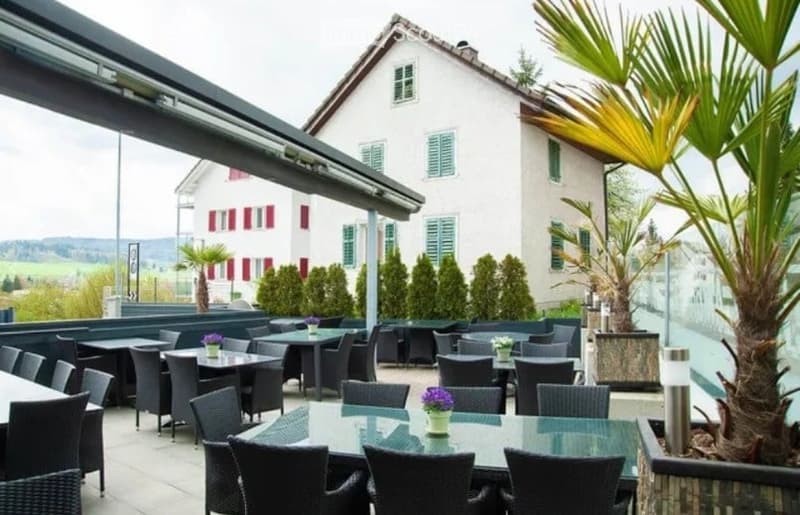 Outdoor seating area with tables and chairs, surrounded by potted plants and greenery, with a view of a traditional house in the background.