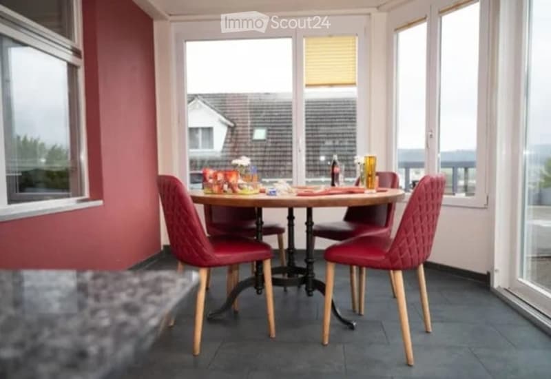 Bright room with large windows offering a view, featuring a round wooden table with four red upholstered chairs, a vase with flowers, and a bottle on the table.
