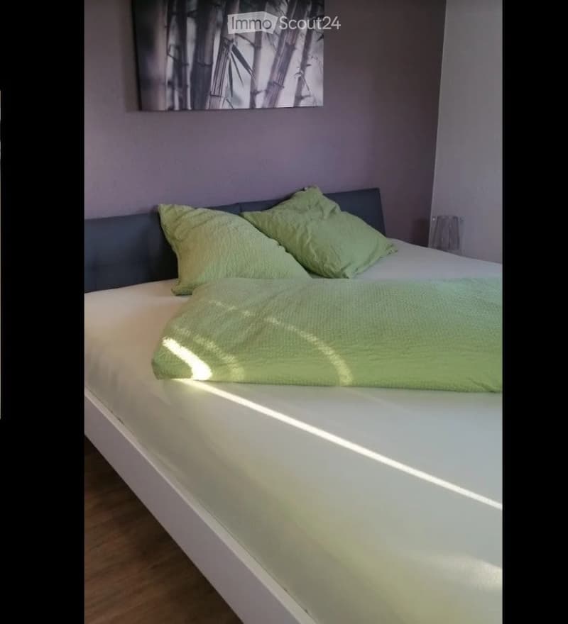 A modern bedroom with a white bed, green pillows and blanket, and a grey headboard. A bamboo-themed artwork hangs on the wall above the bed.