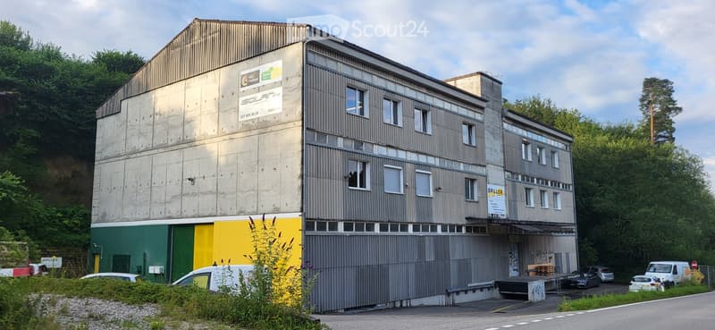 Multi-story industrial building with a mix of concrete and metal siding, featuring signage for 'Solana' and 'Spiller', surrounded by greenery and parked vehicles.