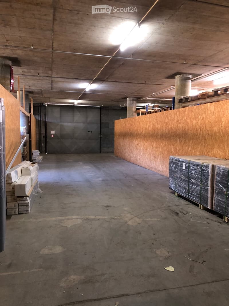 Empty warehouse or storage area with concrete floors, exposed ceiling beams, and stacks of boxes and materials along the walls.