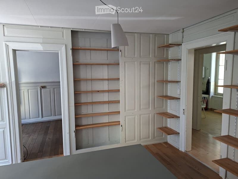 Empty room with wooden flooring, white walls, built-in wooden shelves, and a hanging lamp.