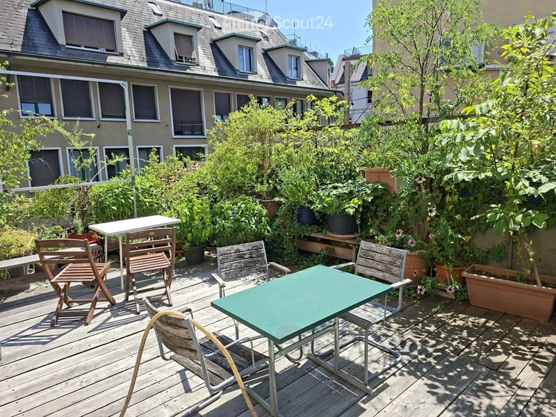 A wooden decked outdoor area with a green table, several chairs, and lush potted plants, surrounded by a multi-story building with multiple windows.