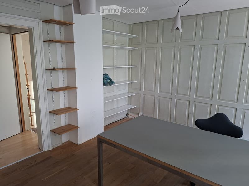 A modern, minimalist room with wooden flooring, white paneled walls, built-in shelves, a grey table, and a black chair.