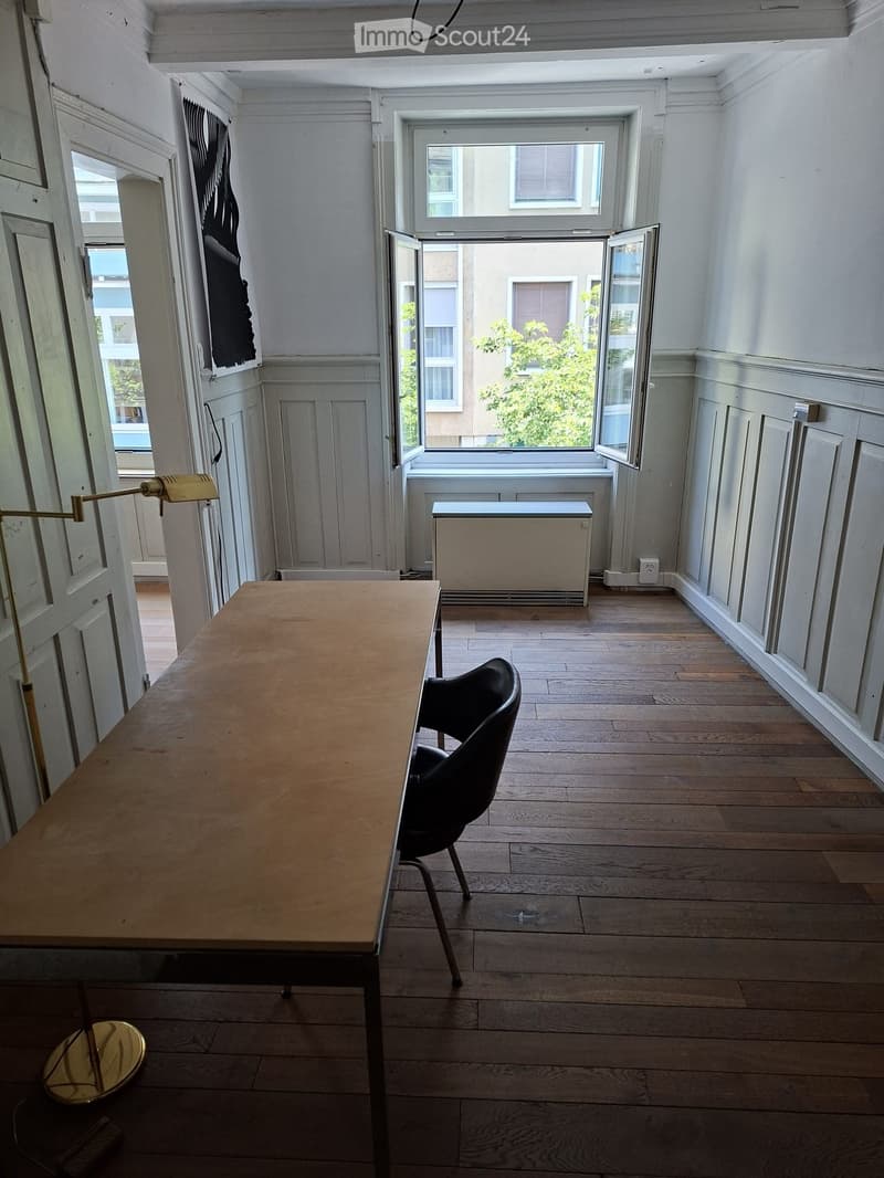 Bright room with wooden flooring, large windows, a table, a chair, and a wall-mounted lamp.