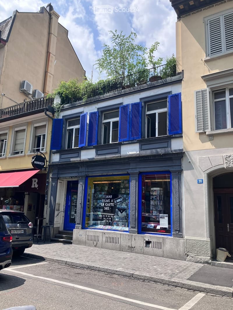 A two-story building with a storefront on the ground floor and residential apartments above, featuring blue shutters and a balcony with greenery.