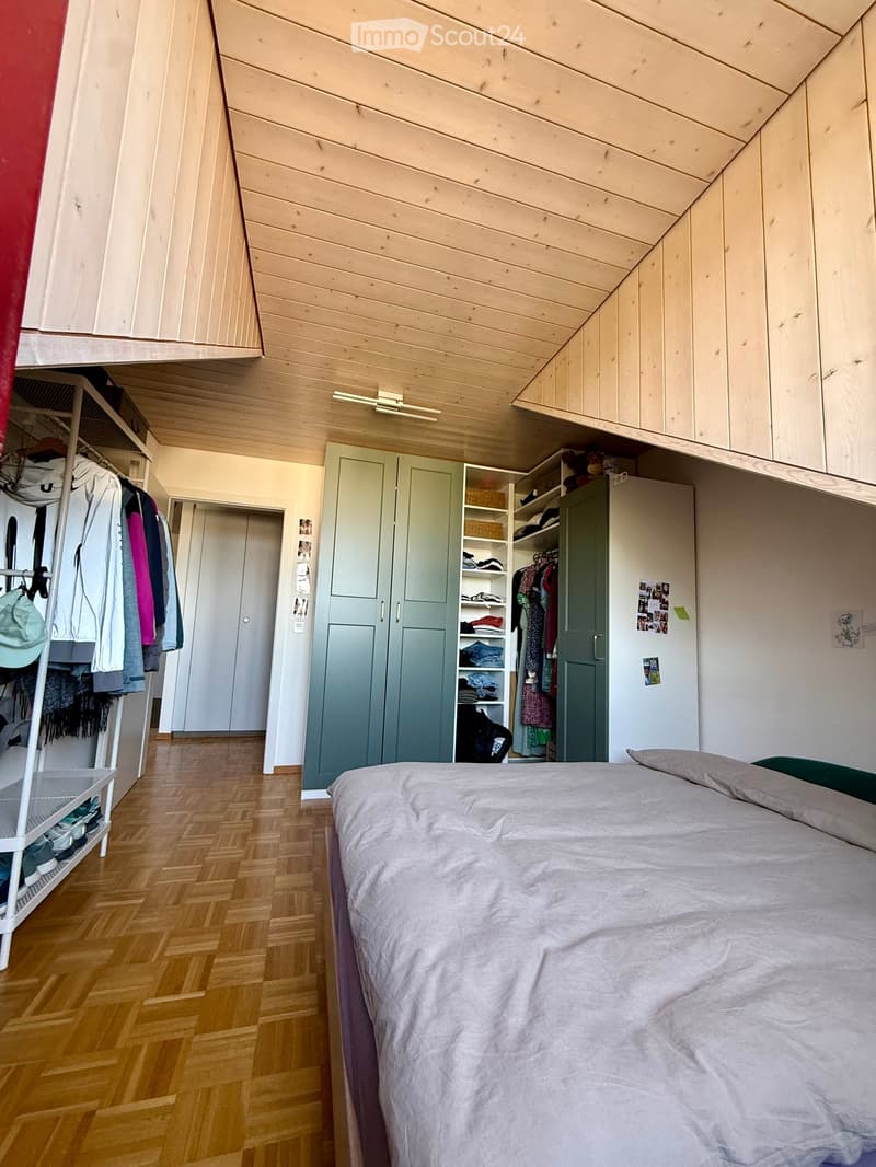 Cozy bedroom with wooden flooring, slanted wooden ceiling, and built-in wardrobes. Features a neatly made bed with pillows and a duvet, along with a clothing rack and shelves.
