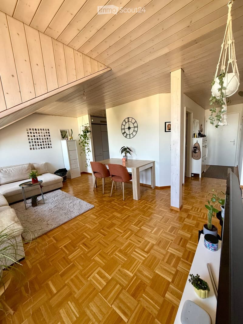Modern living room with wooden herringbone flooring, white walls, and exposed wooden ceiling beams. Features a cream sofa, glass coffee table, dining table with chairs, and decorative plants.