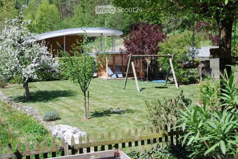 A well-maintained garden with a modern wooden play structure, swings, a round nest swing, and a small seating area with a bench and chairs. The area is surrounded by a wooden fence and lush greenery, including flowering trees and shrubs.