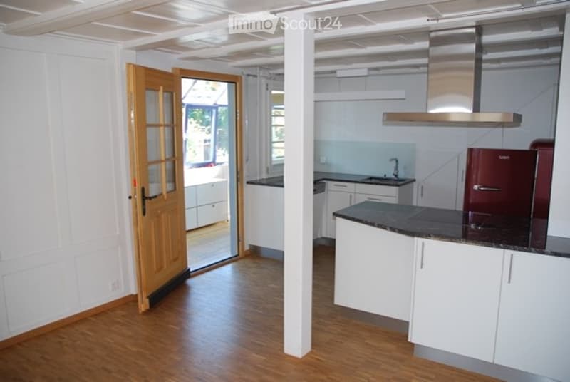 Modern kitchen with white cabinets, dark granite countertop, stainless steel sink and range hood, and a red refrigerator. Wooden flooring and an open doorway leading to another room.