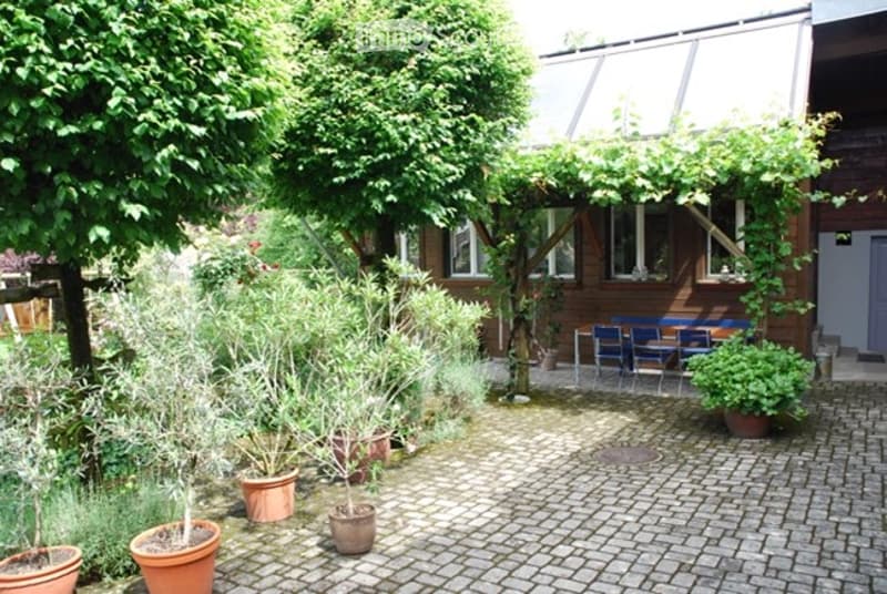 A well-maintained garden area with a brick-paved pathway, several potted plants, and a small seating area with a table and chairs under a pergola.