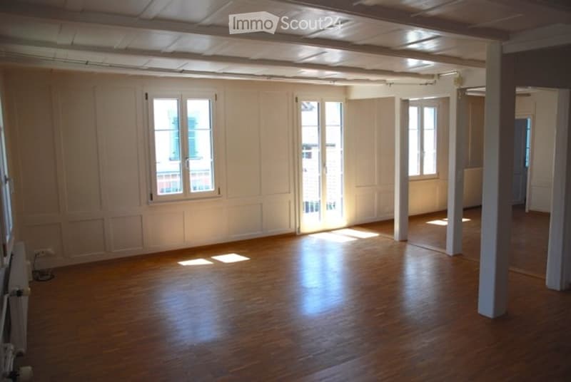 Empty room with wooden flooring, white walls, and large windows allowing natural light. No furniture or equipment present.