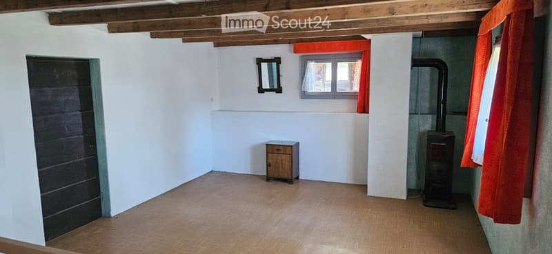 Empty room with wooden flooring, white walls, a small wooden cabinet, a black door, a window with orange curtains, and a small heating stove.