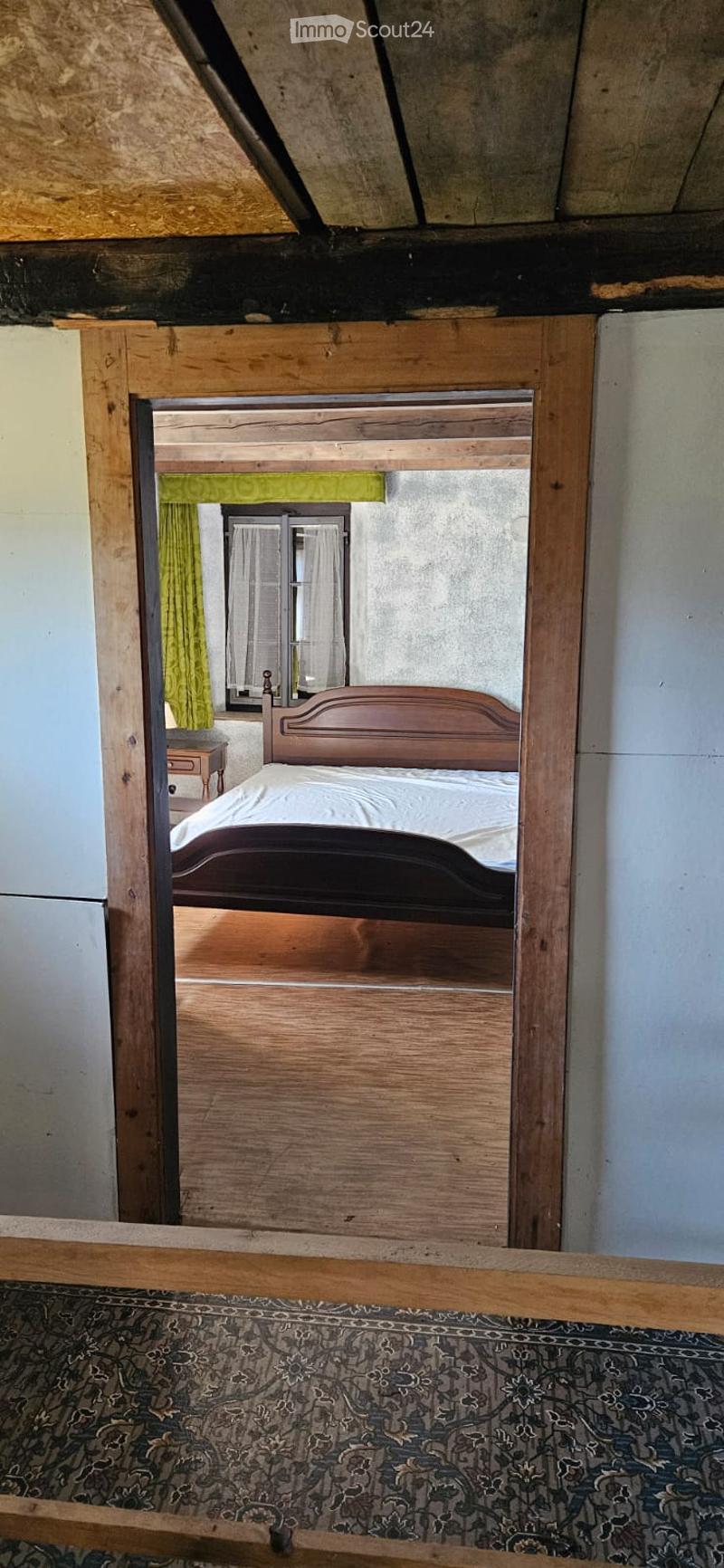 A bedroom with a wooden bed, nightstand, and mirror. The room features patterned carpeting and a window with a green curtain.