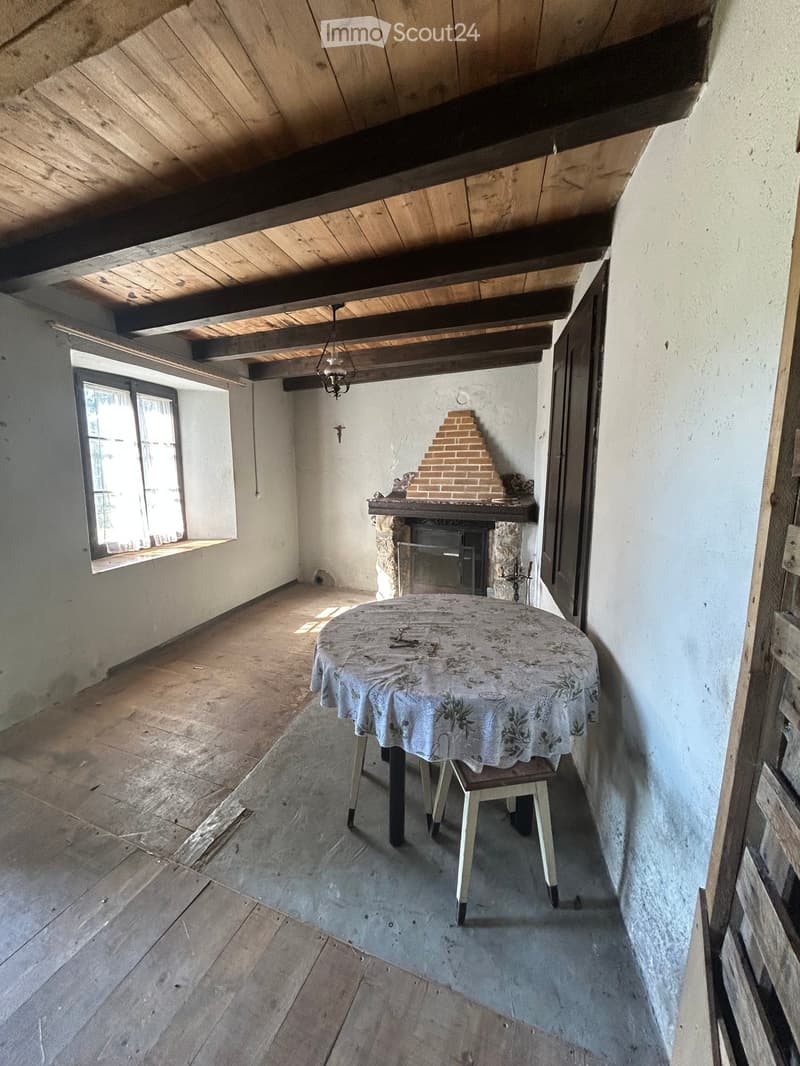 An attic room with wooden beams, a fireplace, a table with a floral tablecloth, and a window letting in natural light.