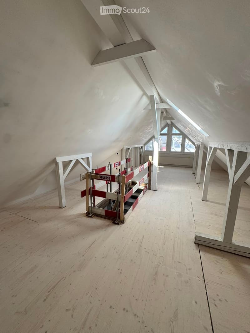 Empty attic space with wooden flooring, exposed wooden beams, and skylights allowing natural light. Construction barriers are present, indicating ongoing work.