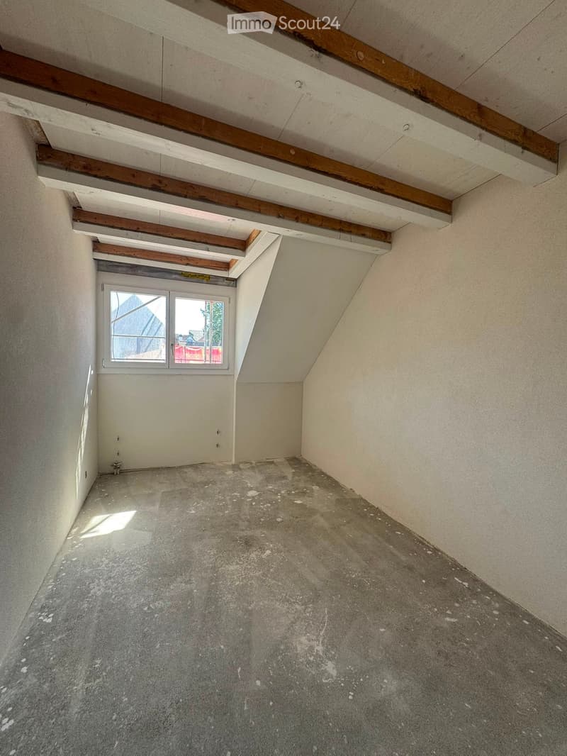 Empty room with exposed wooden beams on the ceiling, concrete floor, and a single window with a view of the outside.