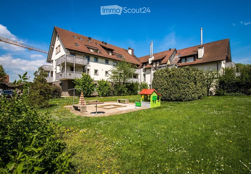A well-maintained garden area with a small playhouse, benches, and a table, surrounded by a two-story residential building with balconies and a red-tiled roof.