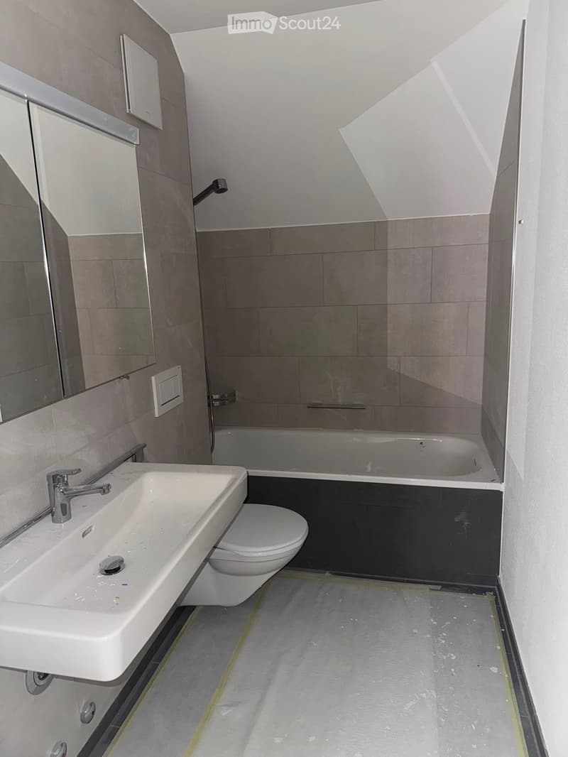 Modern bathroom with a white sink, chrome faucet, toilet, bathtub, and a large mirror above the sink. The walls are tiled in a neutral color scheme, and the floor appears to be under construction or renovation.