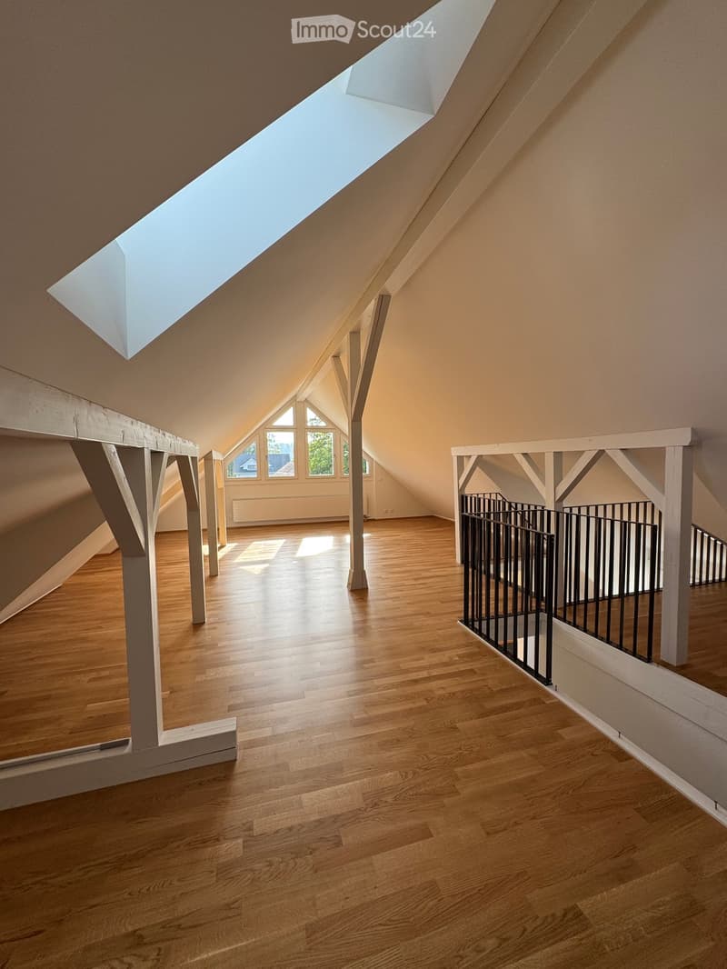Bright, open attic space with wooden flooring, exposed wooden beams, and a skylight. The area includes a staircase leading to another level and several windows letting in natural light.