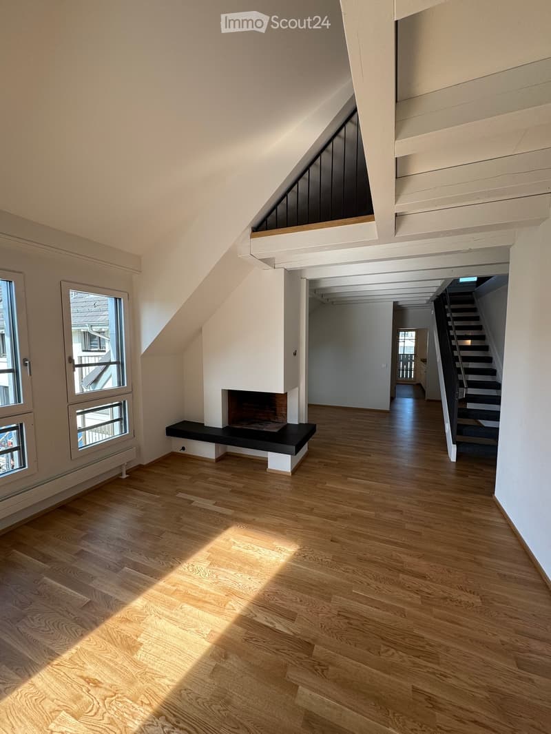 Bright, empty room with wooden flooring, large windows, and a fireplace. The space features a staircase leading to an upper level and a balcony area visible in the background.