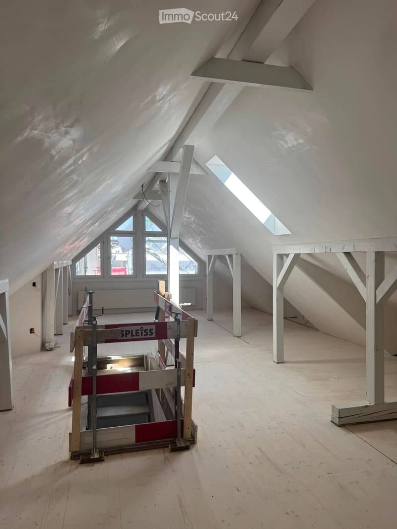 Empty attic space with wooden flooring, exposed beams, and skylights allowing natural light. Construction barriers and tools are present, indicating ongoing renovation.