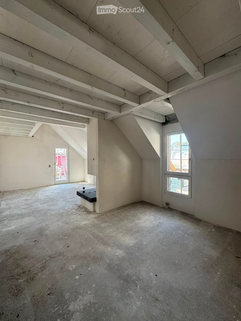 Empty attic space with exposed wooden beams, concrete floor, and large windows allowing natural light. Minimal furnishings include a small black bench and a ladder leading to another area.