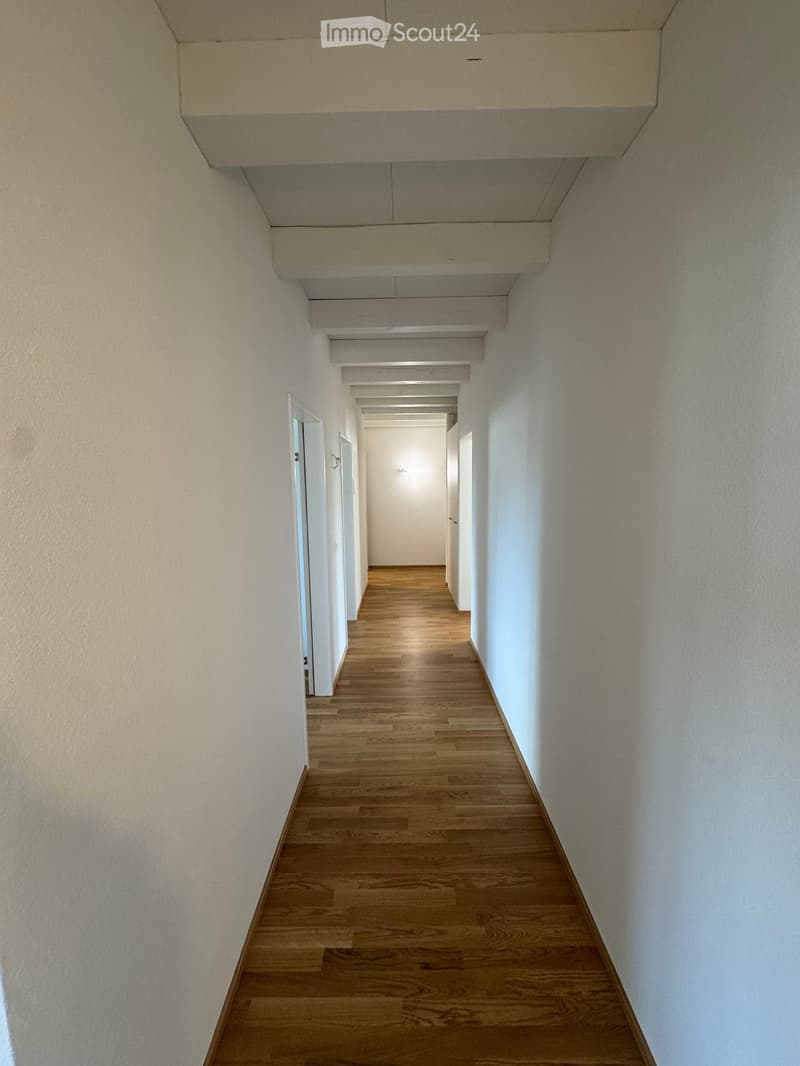 Empty hallway with wooden flooring and white walls, featuring a series of doors and a ceiling with exposed beams.