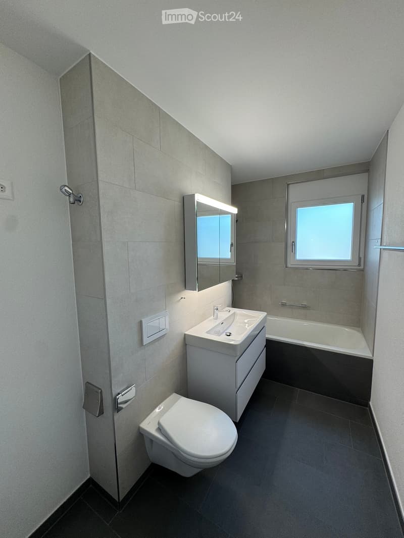 Modern bathroom with a toilet, sink, bathtub, and mirror. The room features grey tiled walls and a window providing natural light.