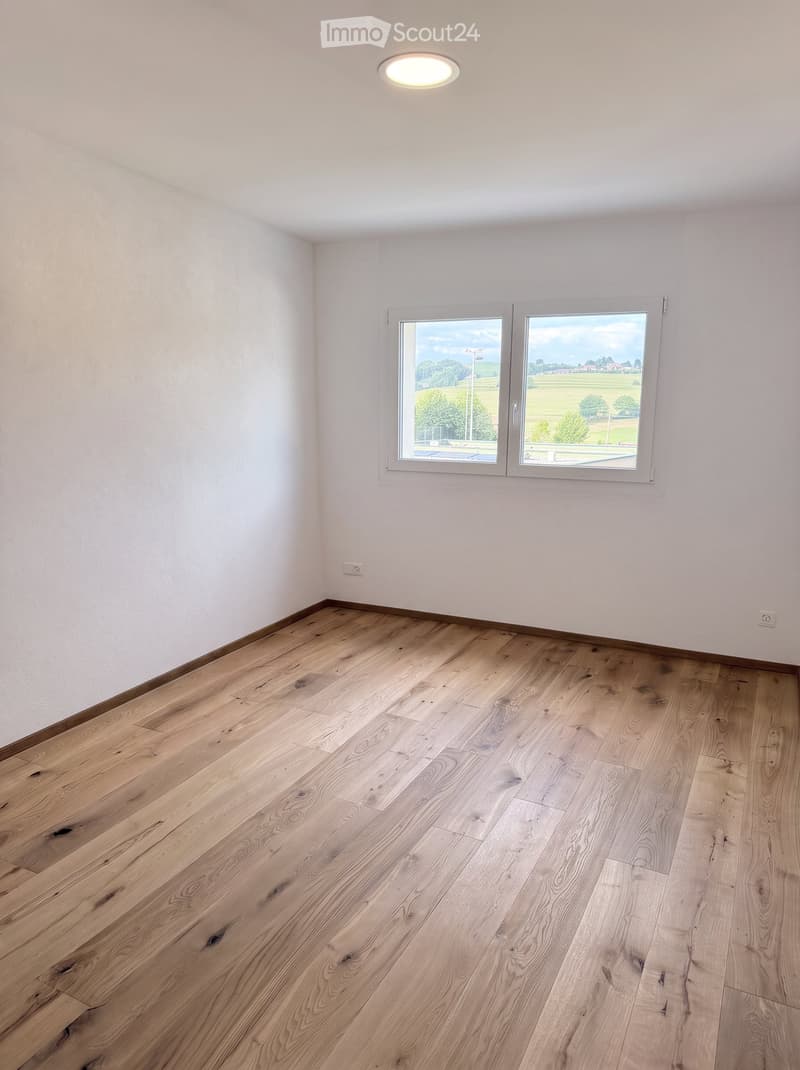 Empty room with light wooden flooring, white walls, and large windows offering a view of a green landscape.