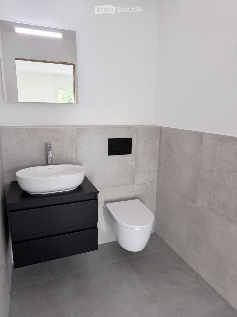 Modern bathroom with a white sink, black cabinet, mirror, and toilet. Grey tiled walls and floor.
