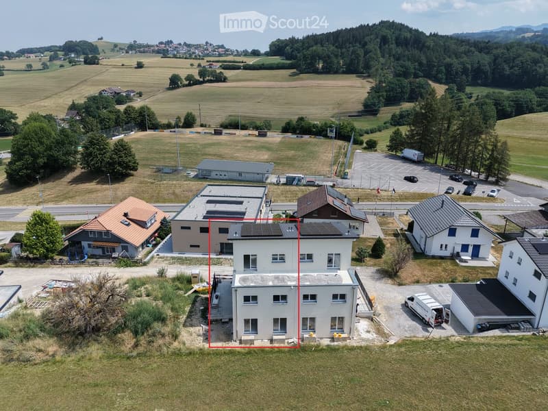 Aerial view of a two-story house with a gray exterior, multiple windows, and a solar panel on the roof, surrounded by a parking area and other buildings in a rural setting.