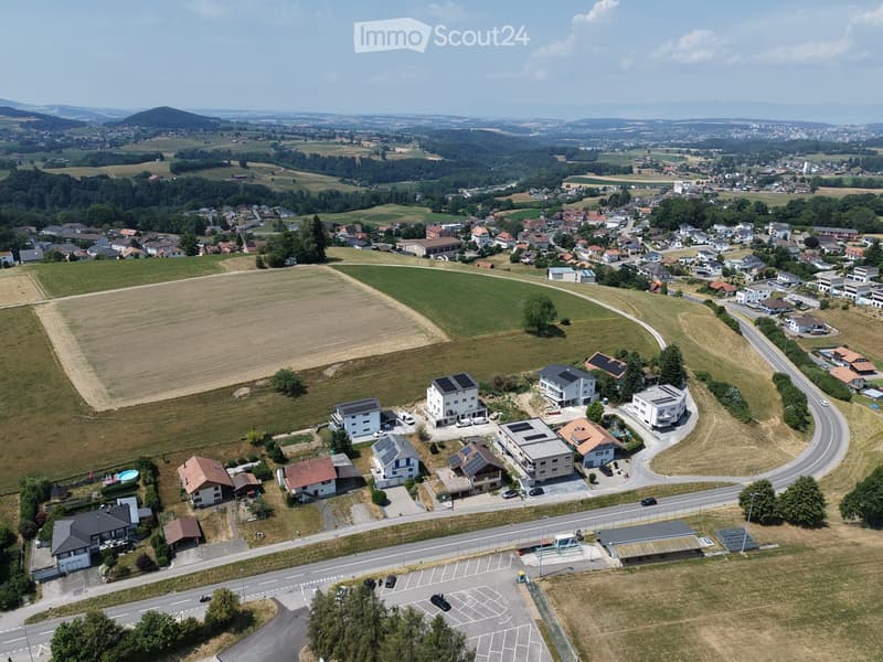 Aerial view of a suburban neighborhood with multiple houses, solar panels, parking lots, and surrounding fields and forests.