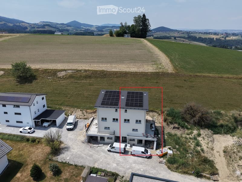Aerial view of a two-story house with solar panels on the roof, surrounded by a rural landscape with fields and trees. The house has a garage, driveway, and several parked vehicles.