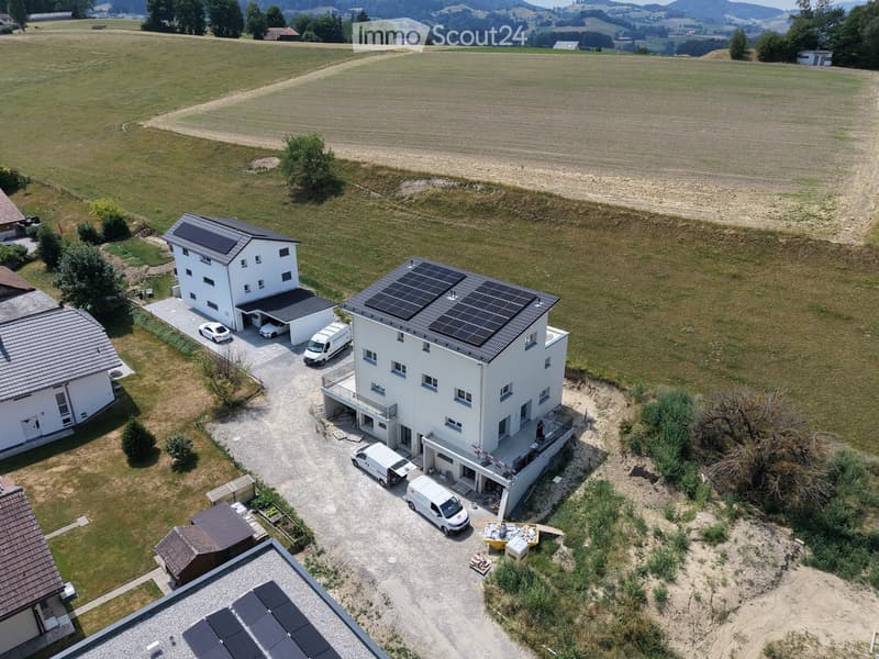 Aerial view of a modern two-story house with solar panels on the roof, surrounded by a rural landscape with fields and trees. The house has a driveway with several parked vehicles, including vans and a car, and a small garden area near the house.