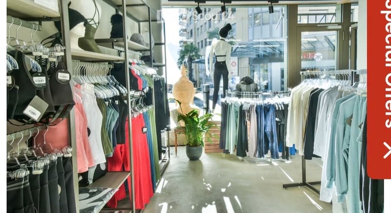 Retail clothing store interior with clothing racks, mannequin, and potted plant. Visible through large windows is an urban street scene with buildings and palm trees.