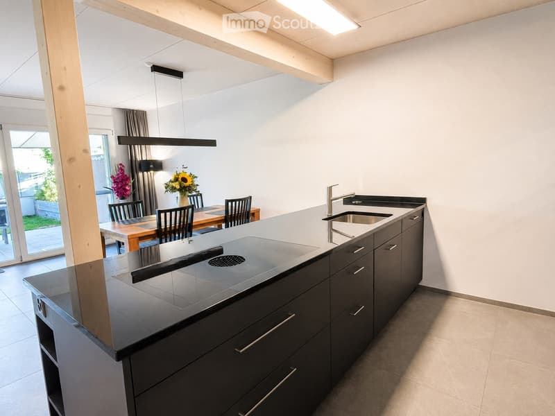 Modern kitchen with dark grey cabinets, a central island with a built-in induction cooktop, a sink, and a dining area with a table and chairs. The space features large windows and a sleek, contemporary design.