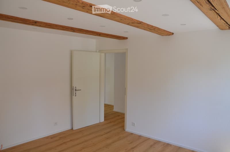 Empty room with light-colored walls, wooden flooring, and exposed wooden beams on the ceiling. Features a closed door and an open doorway leading to another room.