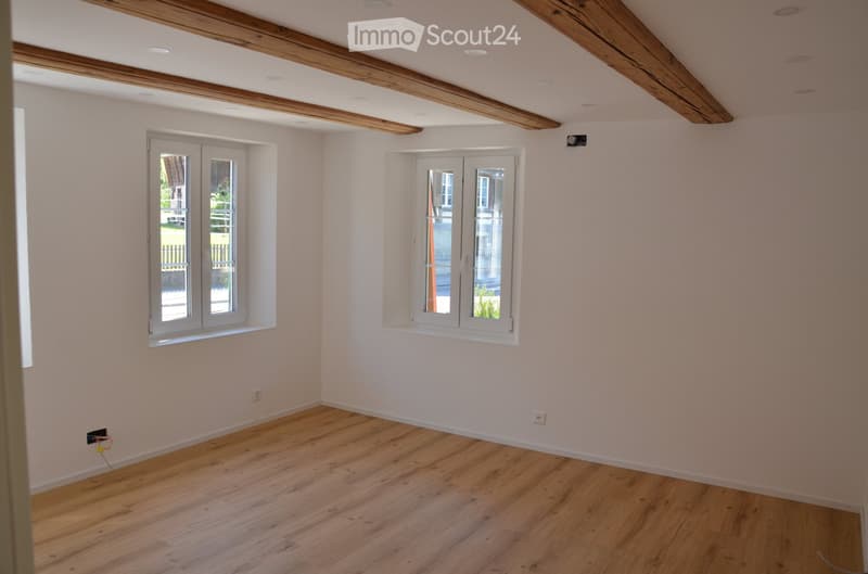 Empty room with light wooden flooring, white walls, exposed wooden beams on the ceiling, and two windows with white frames.