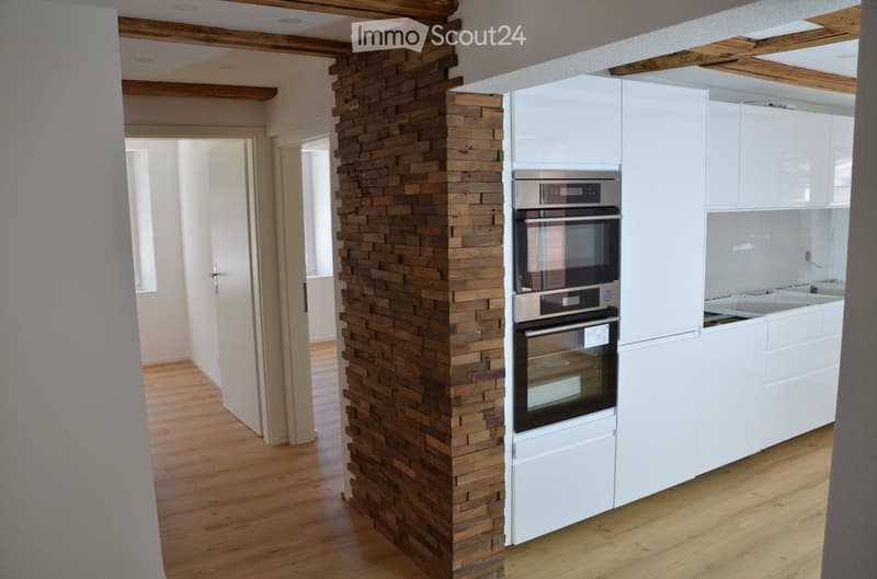 Modern kitchen with white cabinets, a built-in oven, and a wooden accent wall. The floor is made of light wood, and the space is open and bright.