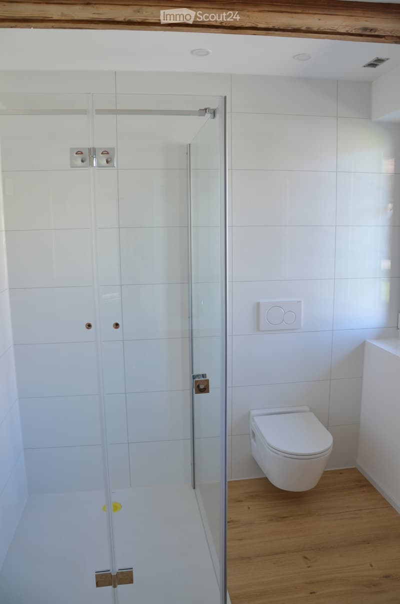 Modern bathroom with white tiled walls, a glass shower enclosure, and a toilet. The floor is wooden, and the ceiling has recessed lighting.