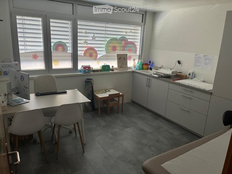 A well-lit room with a desk, chairs, a small table, a sink, and various office supplies. The room features large windows with decorative blinds and a colorful design on the glass.