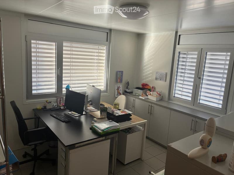 Office space with a desk, computer, chair, and various office supplies. Windows with blinds provide natural light.