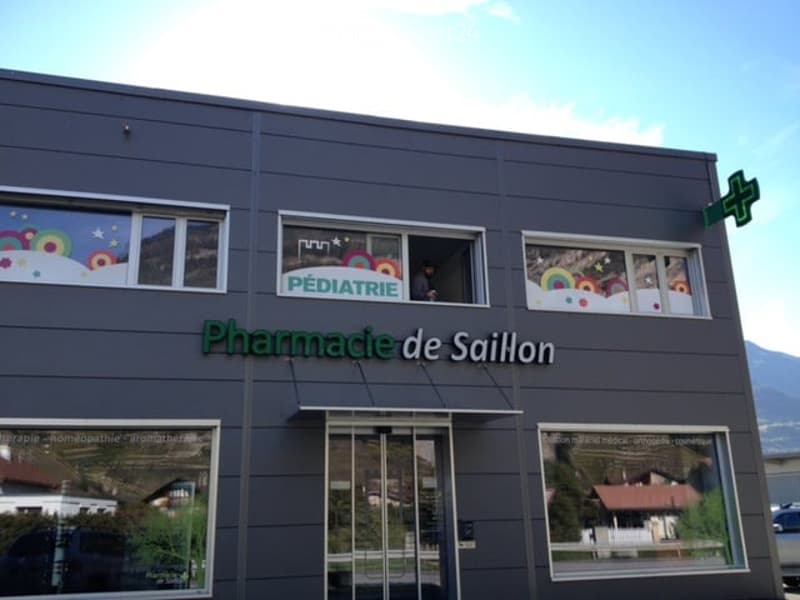 Modern building facade with signage reading "Pharmacie de Saillon" and "Pédiatrie". Windows display colorful children's artwork, and a person is visible in one window. The building has a sleek, dark grey exterior with a small awning above the entrance.