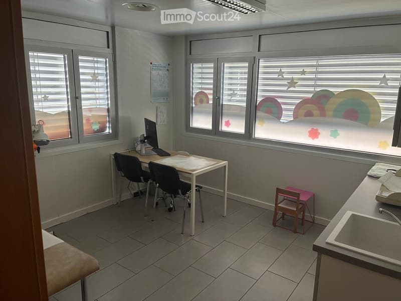 Bright room with large windows, a desk with a computer, two chairs, a small stool, and a sink area.