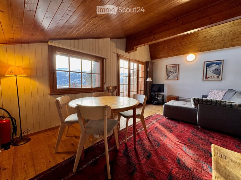 Cozy living room with wooden beams, exposed wooden floor, large window with mountain view, sofa, ottoman, dining table with chairs, television, and artwork on walls.