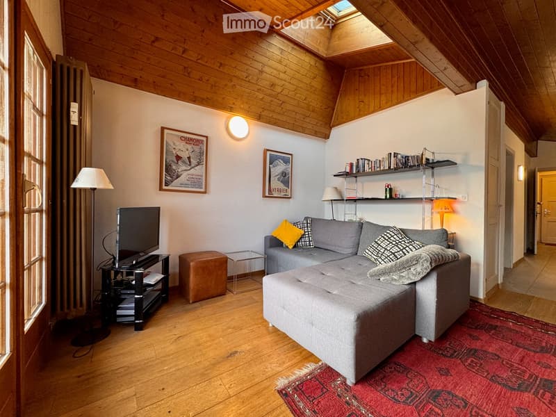 Cozy living room with wooden ceiling, large grey sectional sofa, ottoman, coffee table, TV, floor lamp, shelves, and artwork.