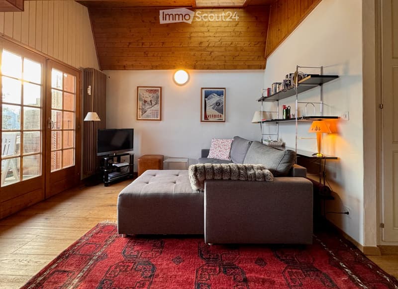 Cozy living room with wooden ceiling, large windows, grey sofa, ottoman, coffee table, television, floor lamp, shelves, and artwork.