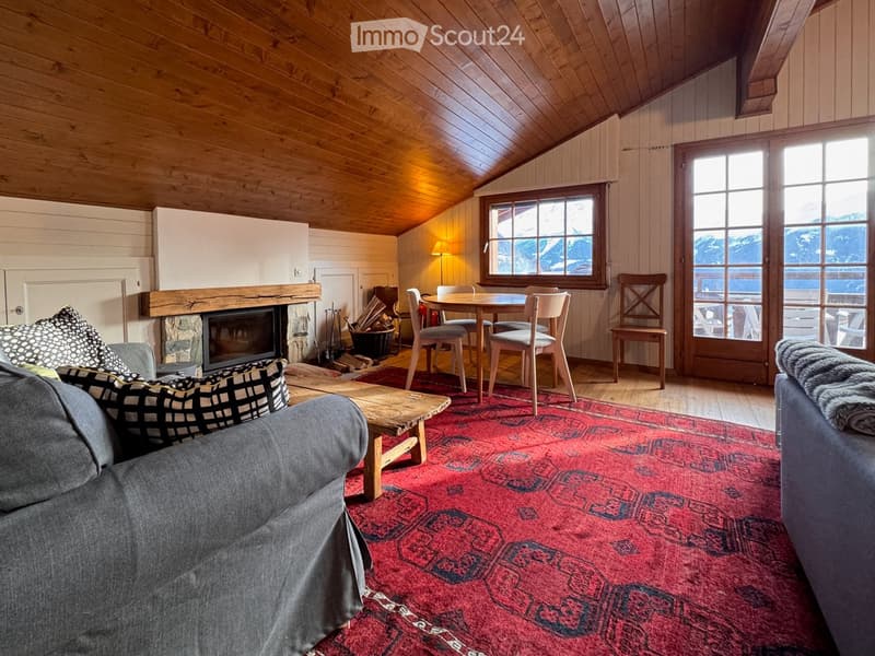 Cozy living room with wooden beams, fireplace, large windows, and a dining area.