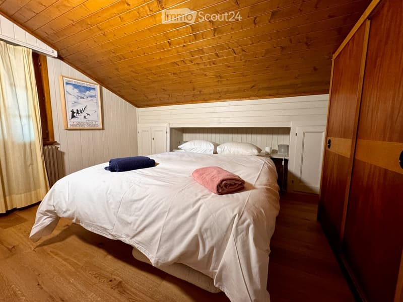 Cozy attic bedroom with wooden beams, a double bed with white linens, two towels, a nightstand, and a framed picture on the wall.